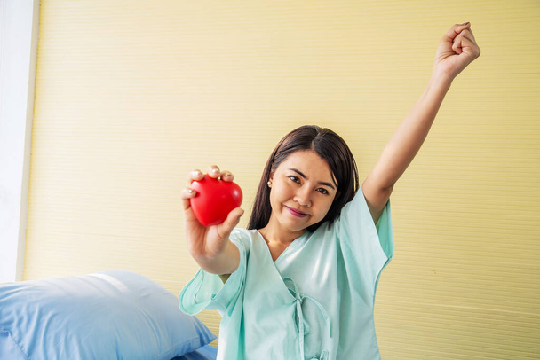 Retrato de mujer joven asiática paciente muestra un corazón rojo en la mano y sentado en la cama con sonrisa y expresiones faciales brillantes, tienen mejores síntomas en la sala de pacientes - Foto, imagen