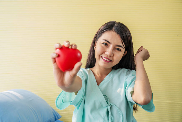 Πορτρέτο της νεαρής γυναίκας ασθενής δείχνει μια κόκκινη καρδιά στο χέρι και κάθεται στο κρεβάτι με χαμόγελο και φωτεινά εκφράσεις του προσώπου, Έχουν καλύτερα συμπτώματα στην πτέρυγα των ασθενών - Φωτογραφία, εικόνα