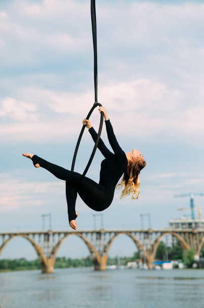 Mulher aerialista executa elementos acrobáticos no aro aéreo pendurado contra fundo de rio, ponte e céu azul
. - Foto, Imagem