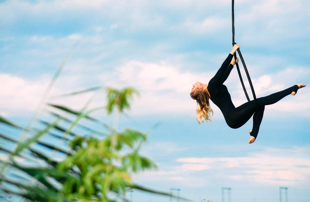 Aerialista mujer realiza elementos acrobáticos en aro aéreo colgante contra el fondo del cielo azul, nubes blancas y hojas. Copiar espacio. - Foto, Imagen