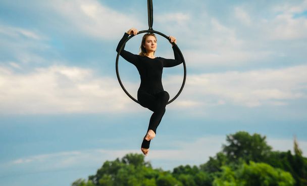 Woman aerialist performs acrobatic elements in hanging aerial hoop against background of blue sky, white clouds and trees. - Photo, Image