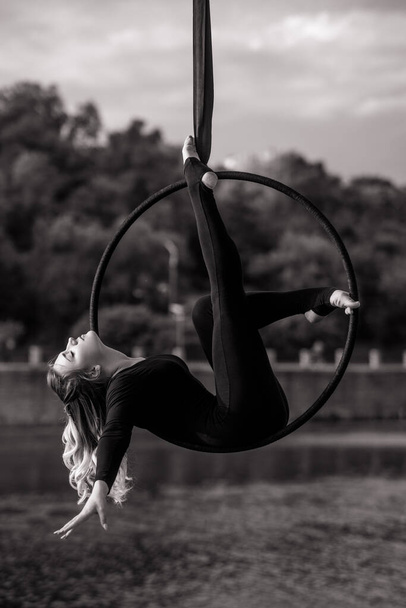 Woman aerialist performs acrobatic elements in hanging aerial hoop against background of river, trees and sky with clouds. Black and white image. - Photo, Image