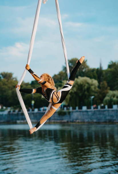 Menina aerialista realiza ginástica dividida em seda aérea pendurada contra fundo de rio, céu azul e árvores
. - Foto, Imagem