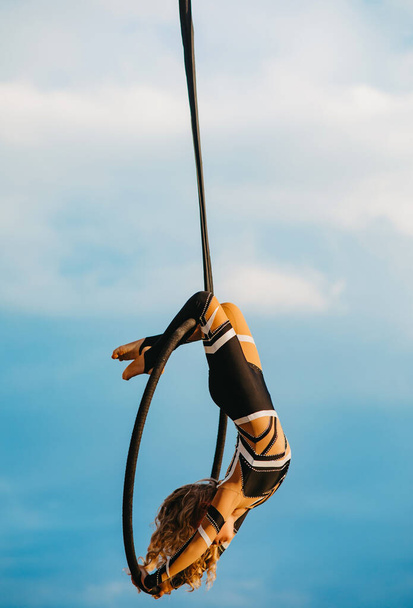 Menina aerialista executa elementos acrobáticos no aro aéreo pendurado contra o fundo do céu azul e nuvens brancas
. - Foto, Imagem