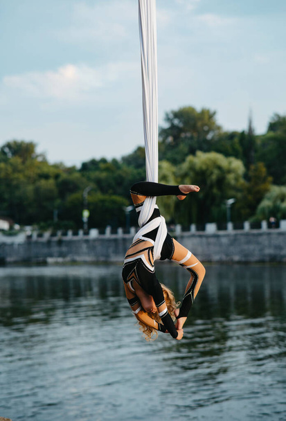 Child girl aerialist performs acrobatic tricks on hanging aerial silk against background of river, sky and trees. - Фото, изображение