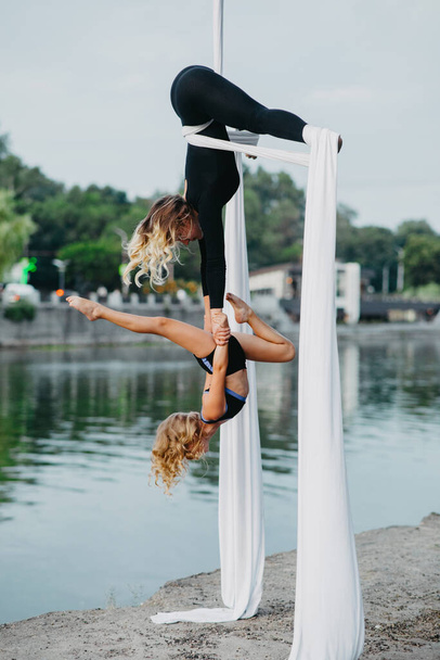 Аэриалисты-женщины и девочки выполняют акробатические трюки на подвешивании воздушного шелка на фоне реки, неба и деревьев. - Фото, изображение