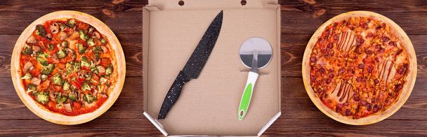 In an open cardboard pizza box is a black kitchen knife and a pizza cutter, next to it are two pizzas with meat and vegetables, on the wooden rustic table, top view - Photo, Image