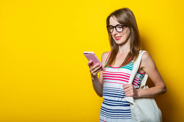 Friendly smiling young girl with glasses looks at the phone, with a linen bag on her shoulder on a yellow background - Photo, image