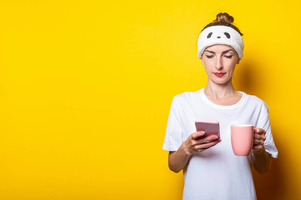 Beautiful young woman with a phone and a cup of coffee on a yellow background. - Fotoğraf, Görsel