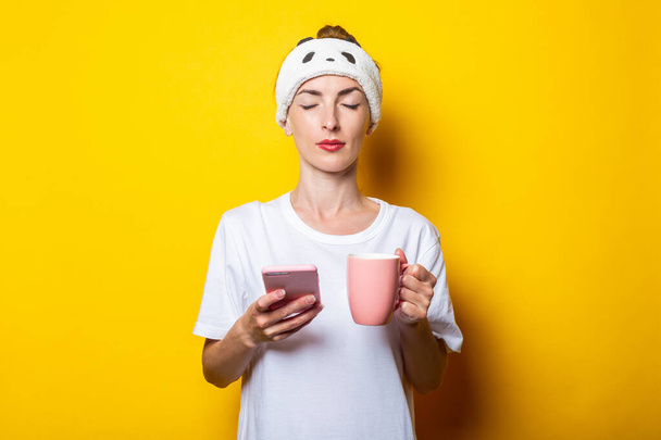 Mujer joven en un arnés de sueño, con los ojos cerrados sostiene un teléfono y con una taza de café sobre un fondo amarillo - Foto, imagen