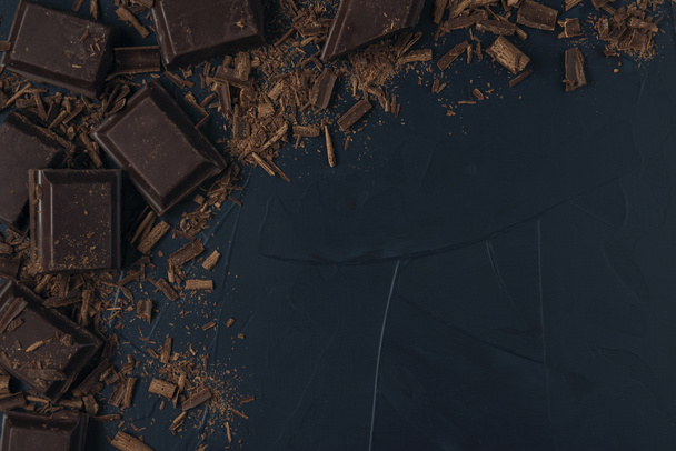 Pezzi di cioccolato con trucioli di cioccolato sullo sfondo strutturato blu scuro. Piatto. Copia spazio. - Foto, immagini