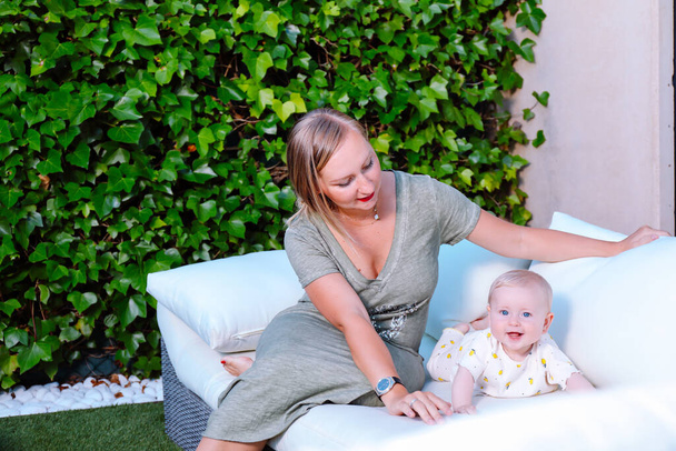 Die junge Mutter sitzt neben ihrem Baby auf dem Sofa im heimischen Garten. Glückliche Familie im Sommerkonzept - Foto, Bild