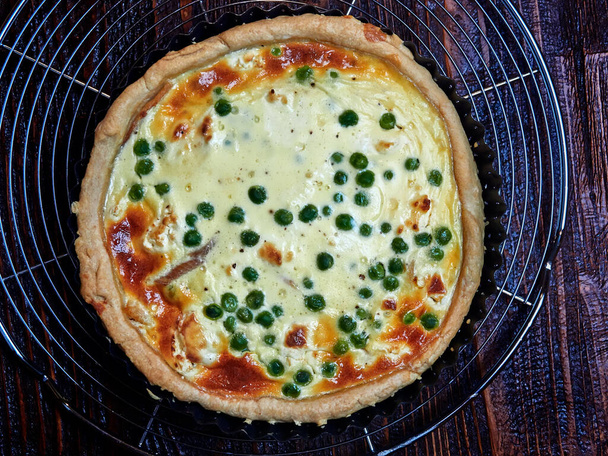 Smoked trout, goat cheese and peas quiche, set on a cooling rack, on a dark  brown wooden board - Photo, image