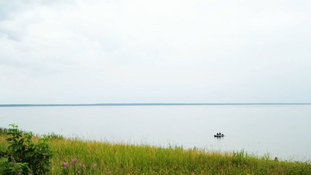 Groen veld en een rustig meer met drijvende rubberboot. Concept. Vers groen gras en bloemen aan de oever van het grote meer op bewolkte lucht achtergrond in de zomer. - Foto, afbeelding