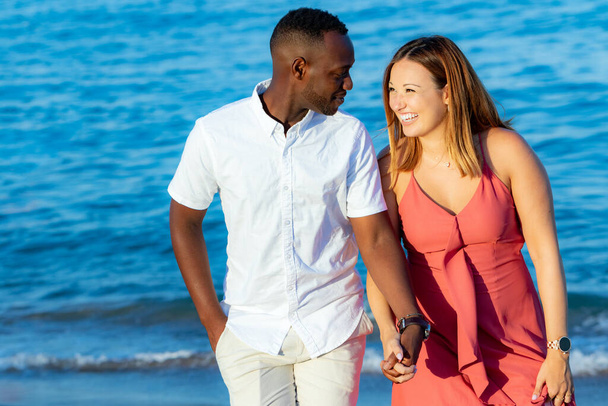 Крупный план портрета молодой разнообразной пары в повседневной одежде, держащейся за руки на пляже. Африканский мужчина и кавказская девушка на синем фоне моря. - Фото, изображение