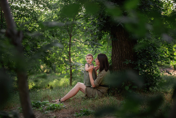 A healthy young mother holds a toddler baby in her arms. A happy family sits on the green grass, under a tall tree, plays, hugs, enjoys a walk in the park. The concept of motherhood, childcare. - Fotografie, Obrázek