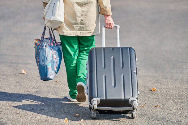 Vieja con maleta sobre ruedas. Mujer con bolsa de equipaje caminando por la calle desde el aeropuerto. Regreso a casa después del viaje. Efecto jet lag. Vista trasera - Foto, imagen