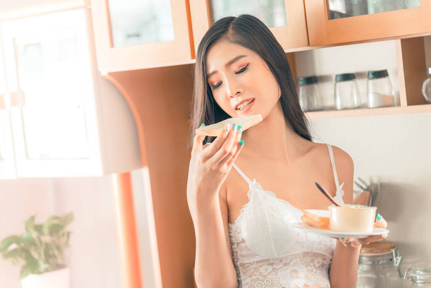 Seductive asian girlfriend in her lingerie is eating bread and butter for breakfast in wooden kitchen. - Fotoğraf, Görsel