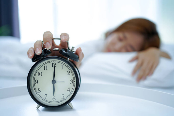 Nuori aasialainen nainen nukkuu sängyssä, käsi painoi torkku painiketta kirkas herätyskello aamulla. Rentouttava konsepti viikonloppuna - Valokuva, kuva