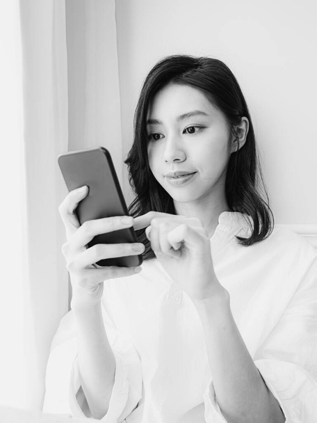 Νεαρή Ασιάτισσα γυναίκα που χρησιμοποιεί smartphone στο σπίτι σε μαύρο και άσπρο - Φωτογραφία, εικόνα