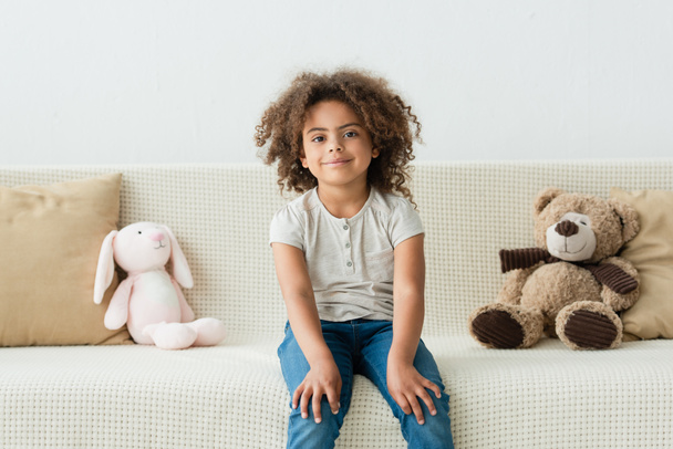 curly african american kid looking at camera and sitting on sofa with soft toys - Foto, Imagem