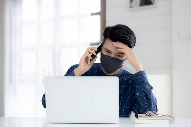 Yüz maskeli Asyalı genç bir iş adamı akıllı telefondan konuşuyor ve bilgisayar başında Coperid-19 'u korumak için çalışıyor. İş adamı karantina işini evden defter ve sosyal mesafeyle yapıyor ve evde kalıyor.. - Fotoğraf, Görsel