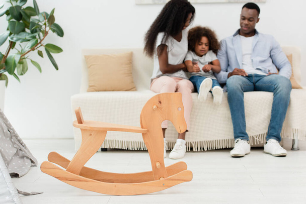 enfoque selectivo de caballo mecedora de madera cerca de la familia afroamericana sentado en el sofá  - Foto, Imagen