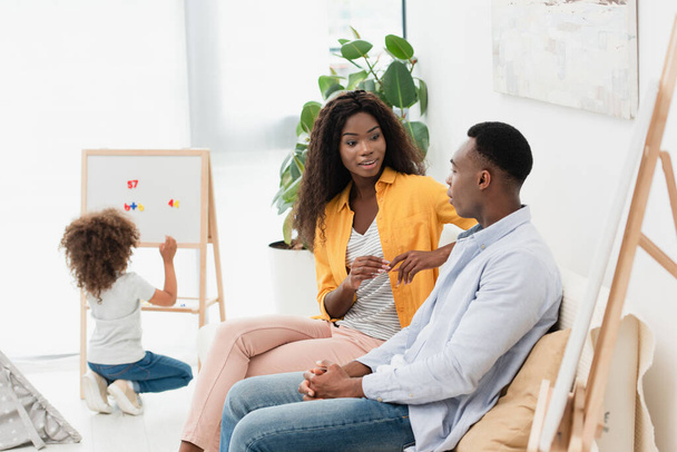 селективное внимание африканских американских родителей, сидящих на диване, в то время как кудрявая дочь сидит рядом с магнитным мольбертом - Фото, изображение