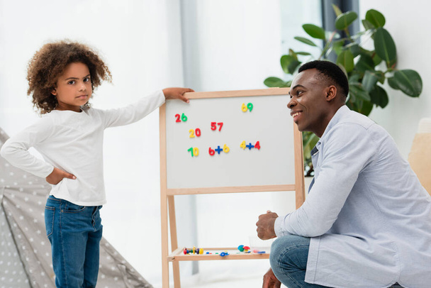 Afrikanisch-amerikanischer Vater schaut ernste Tochter an, die mit der Hand auf Hüfte neben Whiteboard mit Magneten steht  - Foto, Bild