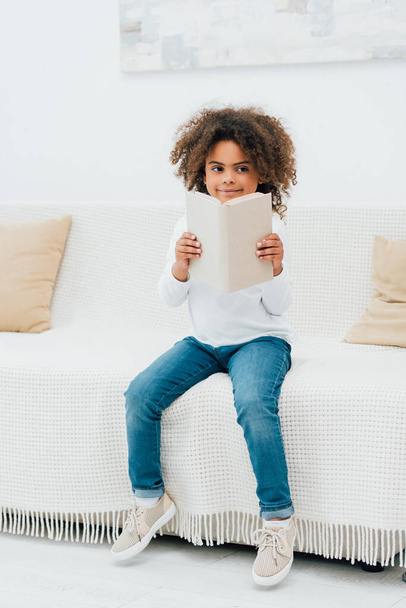 african american kid sitting on sofa and holding book  - Photo, Image