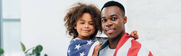 Afro-Amerikan baba ve kızının panoramik konsepti Amerikan bayrağıyla kaplıydı ve kameraya bakıyordu.  - Fotoğraf, Görsel