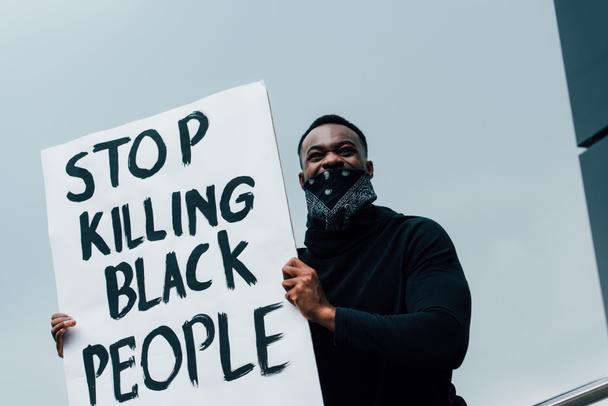 Αφροαμερικάνος άνδρας με μαντήλι στο πρόσωπο κρατώντας πλακάτ και σταματώντας να σκοτώνει μαύρους που γράφουν έξω  - Φωτογραφία, εικόνα