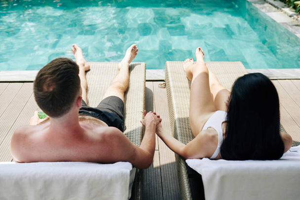 Νεαρό ζευγάρι κρατά το χέρι κατά την ηλιοθεραπεία από την πισίνα και κοιτάζοντας το νερό - Φωτογραφία, εικόνα
