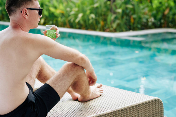 Bonito jovem pensativo sentado à beira da piscina, bebendo bebida fria e olhando para a água - Foto, Imagem