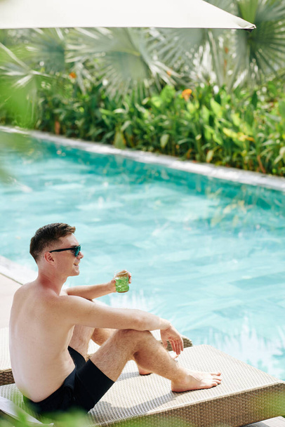 Щасливий молодий чоловік проводить сонячний день у басейні та п'є холодний коктейль
 - Фото, зображення