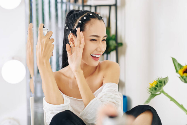 若いアジアの女性は、髪のスプレーを適用する際に目を細めて手で顔を覆う - 写真・画像