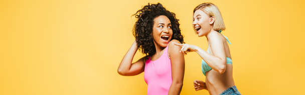imagen horizontal de mujer rubia excitada señalando con el dedo cerca de amigo afroamericano tocando pelo aislado en amarillo - Foto, Imagen