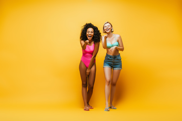 full length view of excited interracial women laughing and pointing with fingers at camera on yellow - Fotografie, Obrázek