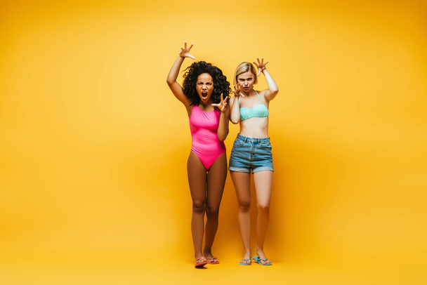 młody międzyrasowe kobiety w lato strój pokazując przerażające gesty patrząc w aparat na żółty - Zdjęcie, obraz
