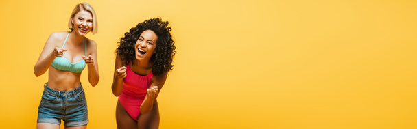 horizontal image of excited interracial women laughing and pointing with fingers at camera isolated on yellow - Фото, зображення