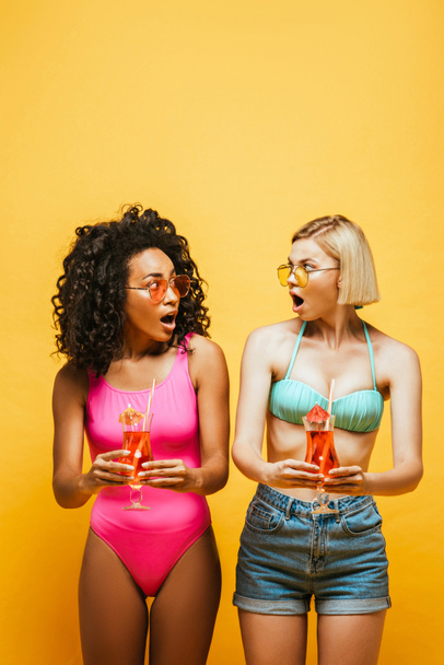 shocked interracial friends in summer outfit holding cocktails and looking at each other on yellow - Фото, зображення