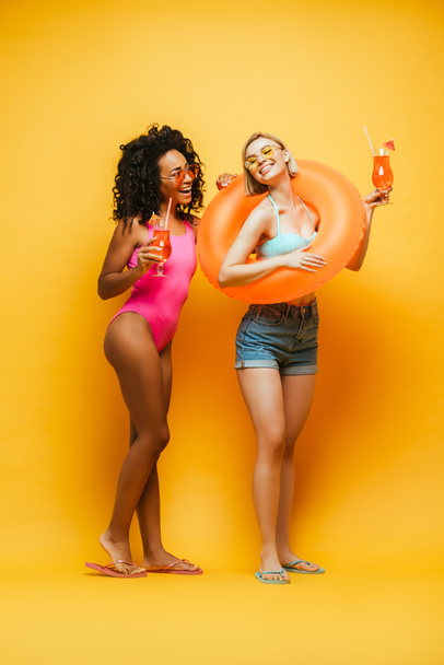 young african american woman in swimsuit looking at blonde friend with swim ring on yellow - Fotó, kép