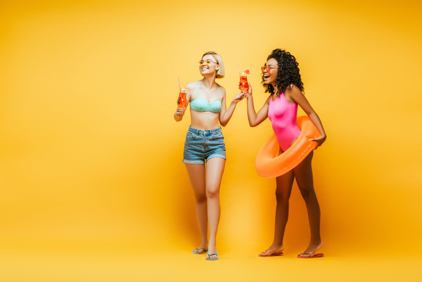 blonde woman with african american friend holding cocktail glasses and swim ring laughing while looking away on yellow - Photo, Image