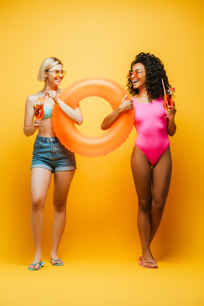 young interracial women in summer outfit holding swim ring and cocktail glasses while looking at each other on yellow - Foto, afbeelding
