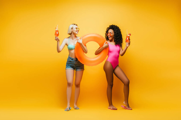 young interracial women in summer outfit posing with swim ring and cocktail glasses while looking at each other on yellow - Foto, imagen