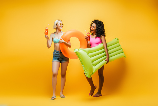 full length view of young multicultural women in summer outfit holding inflatable mattress, swim ring and cocktails on yellow - Φωτογραφία, εικόνα