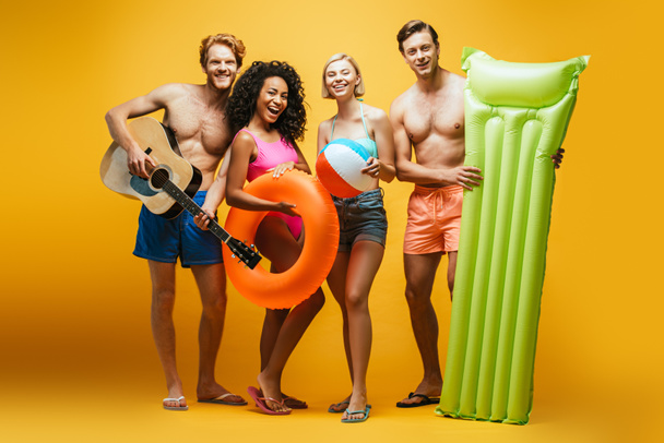 full length view of excited multicultural friends with guitar, inflatable mattress, ball and swim ring looking at camera on yellow - Photo, Image