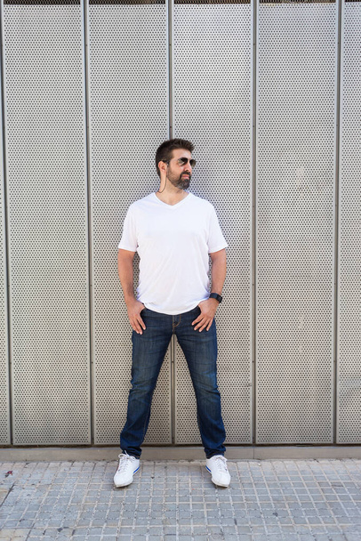 Bearded guy in sunglasses leaning on metallic wall with hands on pocket while looking away - Photo, Image