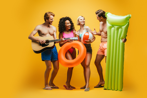 full length view of excited multicultural friends with guitar, inflatable mattress, ball and swim ring talking on yellow - Fotografie, Obrázek