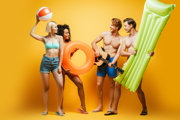 vue pleine longueur de jeunes amis multiculturels en tenue d'été avec guitare, matelas gonflable, ballon et anneau de natation se regardant sur jaune - Photo, image